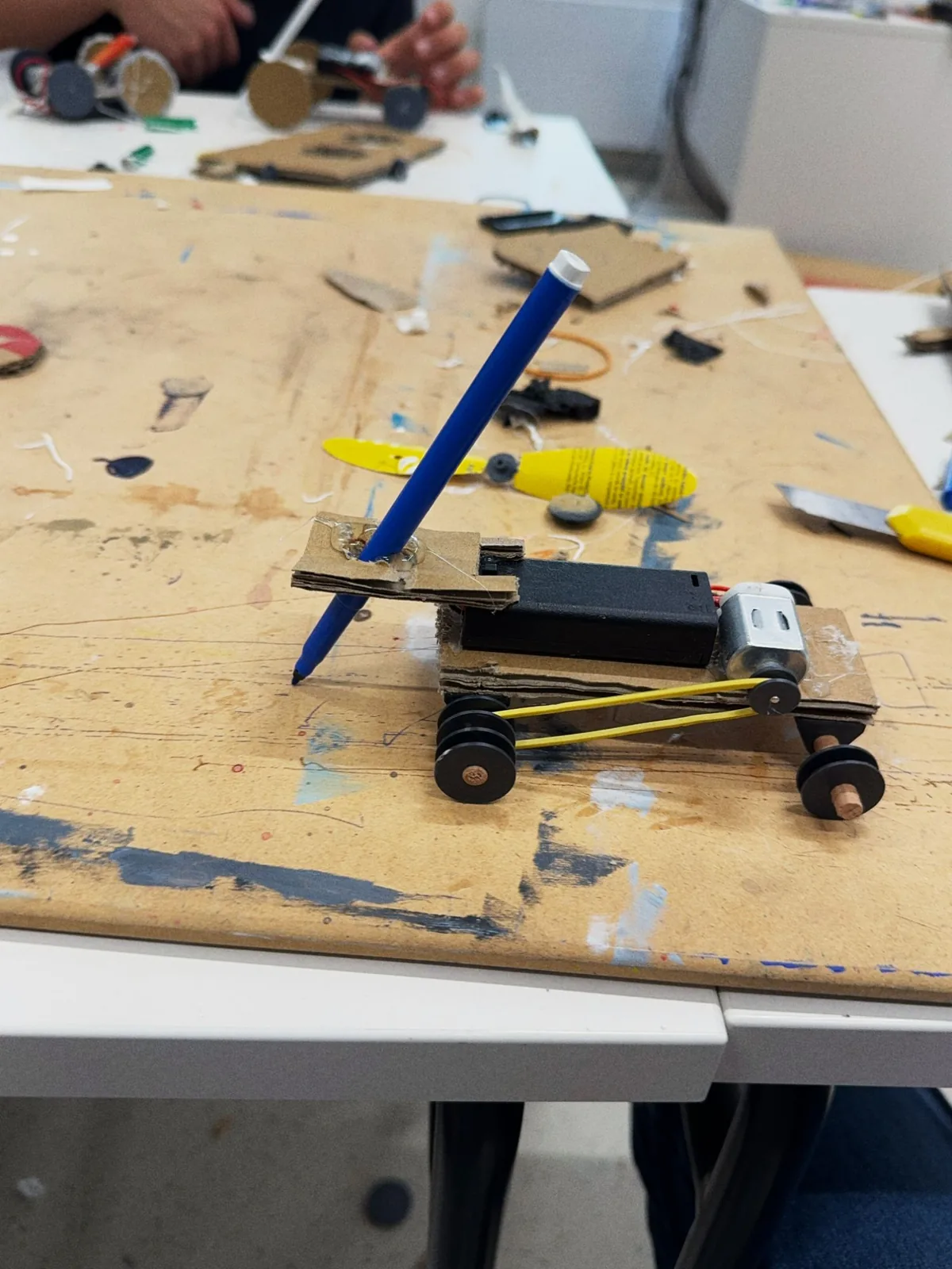 A cardboard car with a blue pencil mounted upright, a DC motor and a rubber band drive